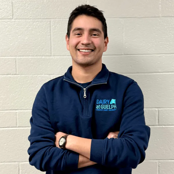 Members A man smiling with his arms crossed, wearing a navy blue quarter-zip pullover with a "Dairy at Guelph" logo, posing against a white brick wall.