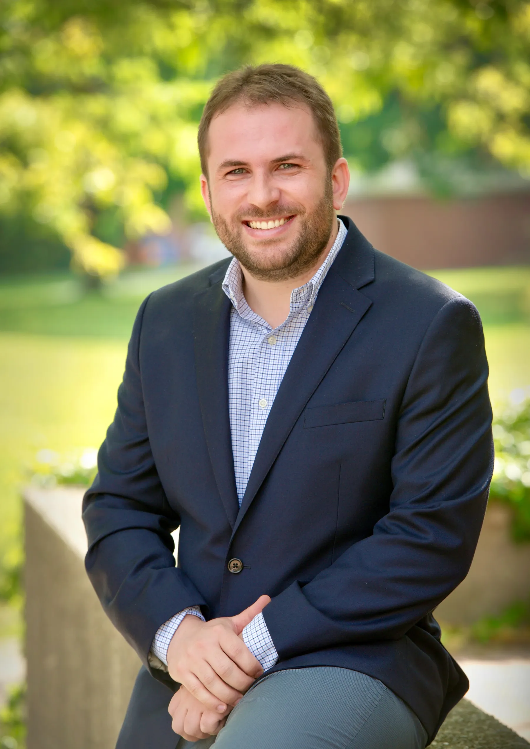 Professional portrait of Dr. Ribeiro smiling, wearing a navy blue blazer and a checkered shirt, with a blurred outdoor greenery background.