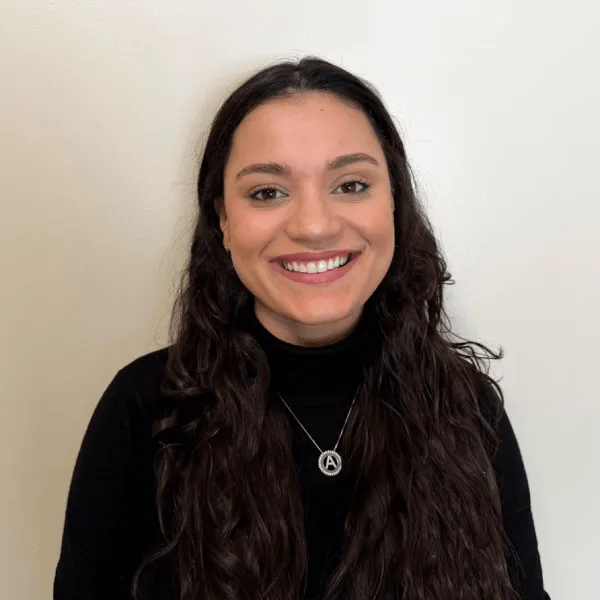 Members A portrait of a smiling woman with long, dark wavy hair, wearing a black turtleneck sweater and a silver initial necklace against a plain off-white background.