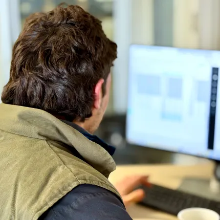 Home Back view of a person with wavy brown hair, wearing a green vest over a dark shirt, sitting at a desk and looking at a computer screen in a blurred office setting.