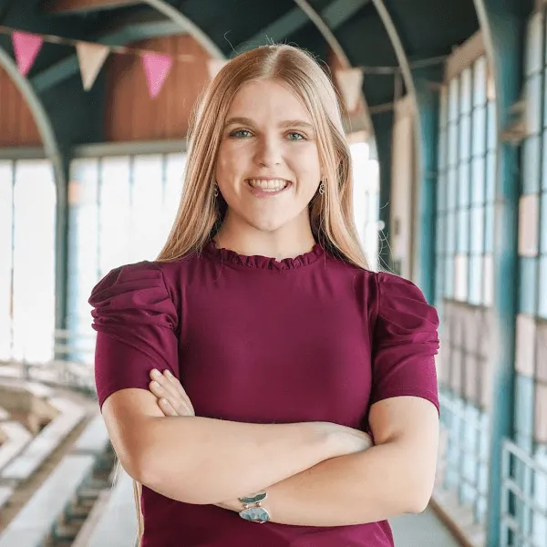 Members A smiling woman with long blonde hair, posing with her arms crossed and wearing a burgundy short-sleeved blouse in an airy, industrial-style building.