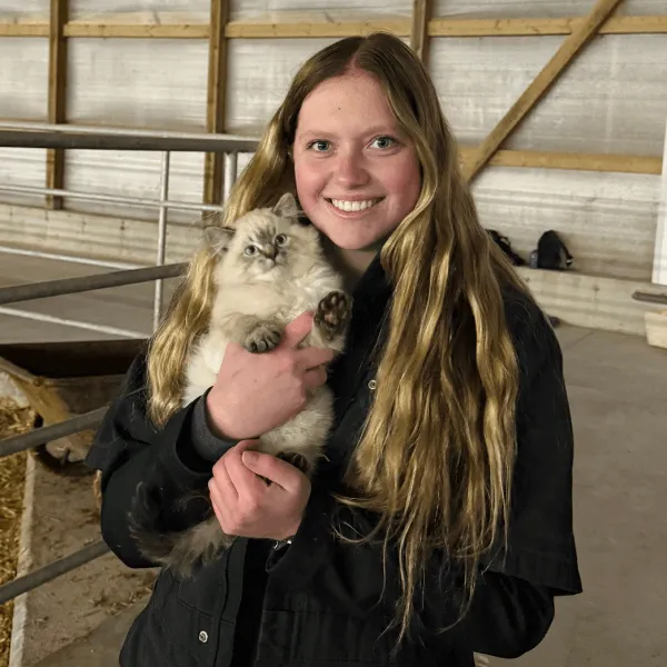 Members A smiling young woman with long blonde hair holding a fluffy grey and white kitten, wearing a black work jacket inside a farm building.