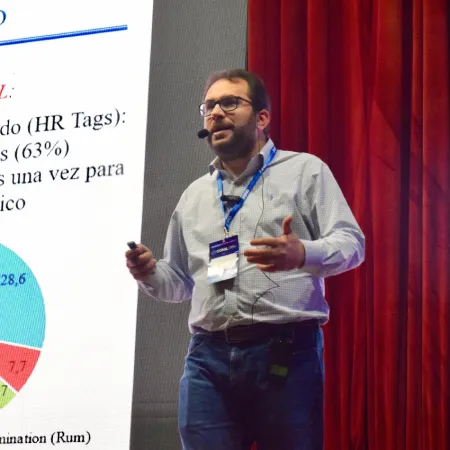Home Dr. Ribeiro stands on a stage, gesturing while giving a presentation. He is wearing a light-colored button-down shirt, jeans, a conference lanyard, and a headset microphone. To his left, a large screen displays a presentation slide with text and a colorful pie chart. Red curtains are visible in the background.