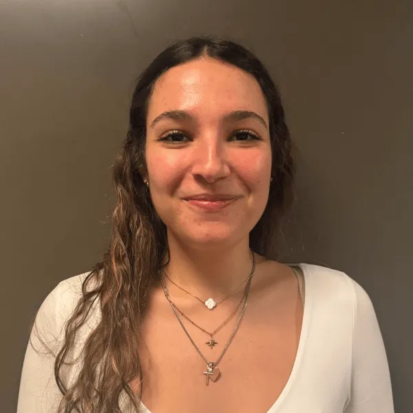 Members A portrait of a smiling woman with long curly brown hair, wearing a white long-sleeved top and layered gold necklaces against a dark grey background.