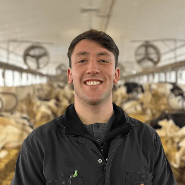 Members A portrait of a man smiling with short dark hair, wearing a black work jacket over a grey shirt. He is standing in a large dairy barn with rows of cows and industrial fans visible in the blurred background.