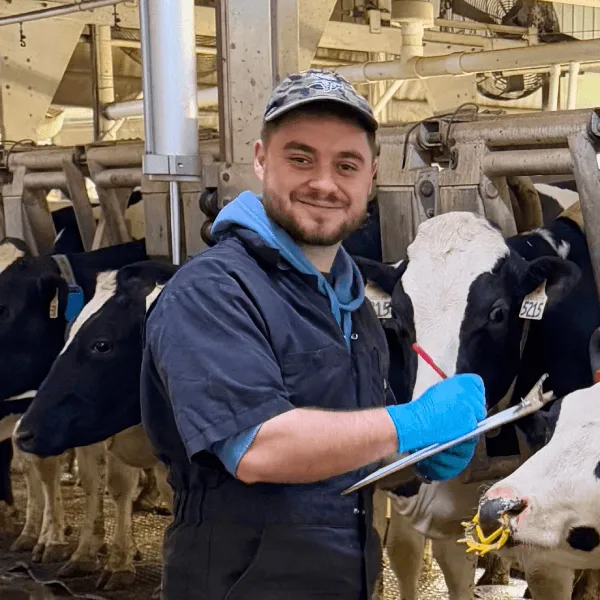 Members A smiling man wearing a camouflage cap, blue hoodie, and black overalls, holding a clipboard and pen while standing next to Holstein cows in a milking parlor.