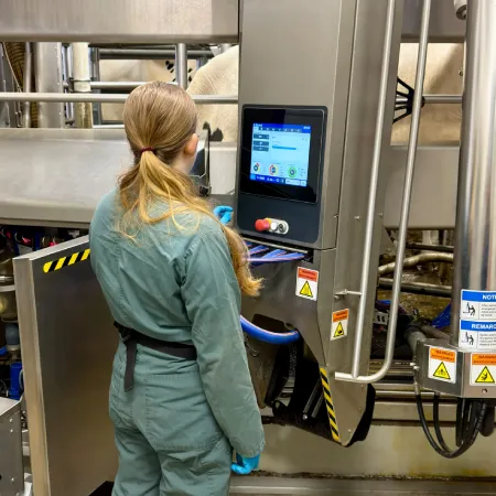 Home A student with long blonde hair tied in a ponytail, wearing green coveralls and blue gloves, stands in a dairy facility. She is looking at a digital control panel mounted on a piece of stainless steel machinery. In the background, the side of a cow is partially visible through the metal railings of the stall.