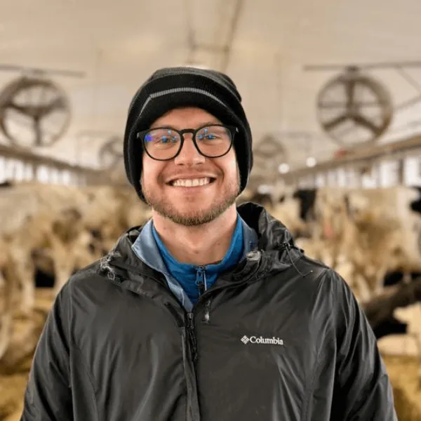 Members A portrait of a smiling man with glasses and a black beanie, wearing a black Columbia jacket inside a dairy facility with cattle in the background.