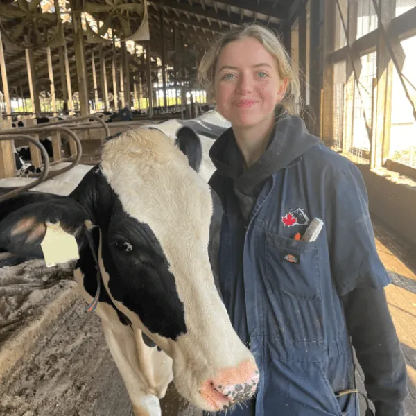 Members A woman smiling while standing next to a Holstein cow inside a barn, wearing navy blue coveralls with a red maple leaf logo.