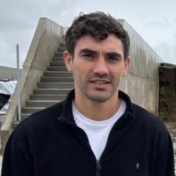 Members A portrait of a man with short, curly brown hair and light facial hair, wearing a black zip-up jacket over a white t-shirt. He is looking at the camera. He is outdoors in a farm setting, with a concrete bunker silo containing dark feed and concrete stairs in the background under a cloudy sky.