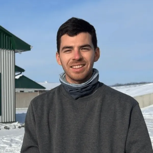 Members A portrait of a man smiling with short dark hair, wearing a grey sweatshirt and a grey neck gaiter. He is standing outdoors in a snowy landscape with a clear blue sky and a farm building with a green roof in the background.