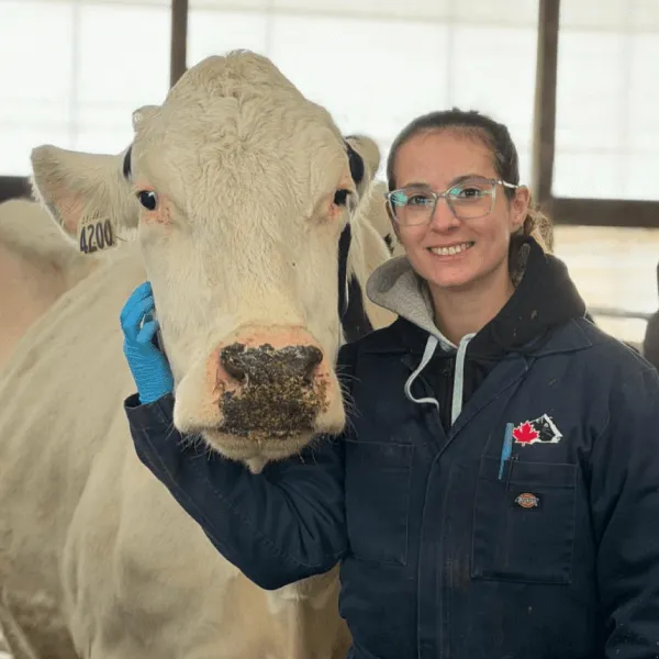 Members A portrait of a woman smiling with glasses, wearing a navy blue work jumpsuit with a Canadian flag patch over a grey hoodie. She has one gloved hand resting on the side of a large white cow's face. They are inside a bright barn with a white tarp wall in the background.