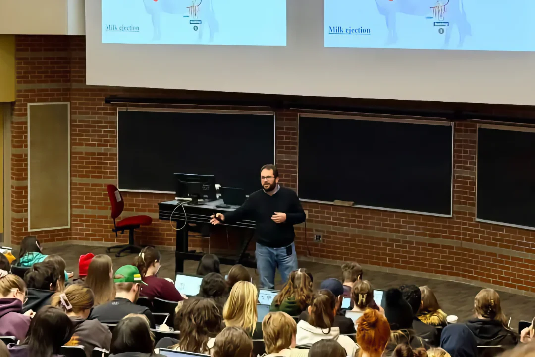 Teaching Wide shot of Dr. Ribeiro standing at the front of a classroom with brick walls and blackboards. He is gesturing during a lecture about animal science, with a diagram of a cow visible on the overhead screens and a large group of students in the foreground.