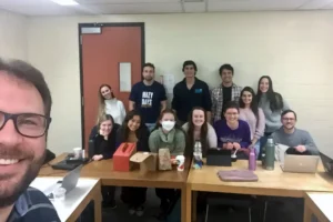Dr. Ribeiro taking a selfie with a diverse group of graduate students from the ANSC 6070 course, gathered around tables in a university classroom during a casual break.