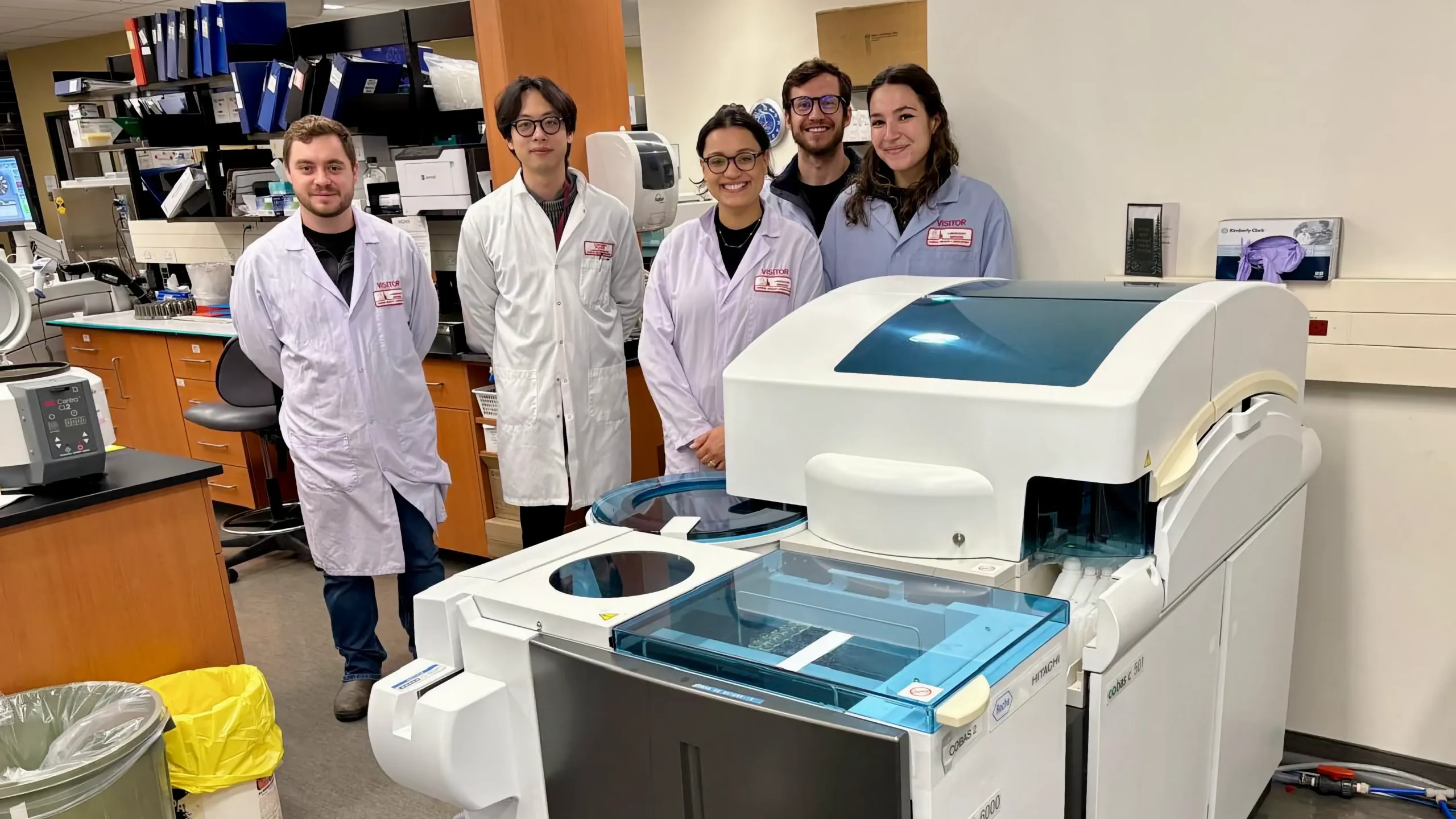 Five researchers wearing white lab coats and visitor badges stand behind a Cobas clinical chemistry analyzer during a site visit to the Animal Health Lab.