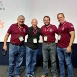 Outreach Four researchers wearing matching maroon polo shirts with event lanyards posing together on a stage in front of a presentation screen at the Novos Enfoques 2026 conference.