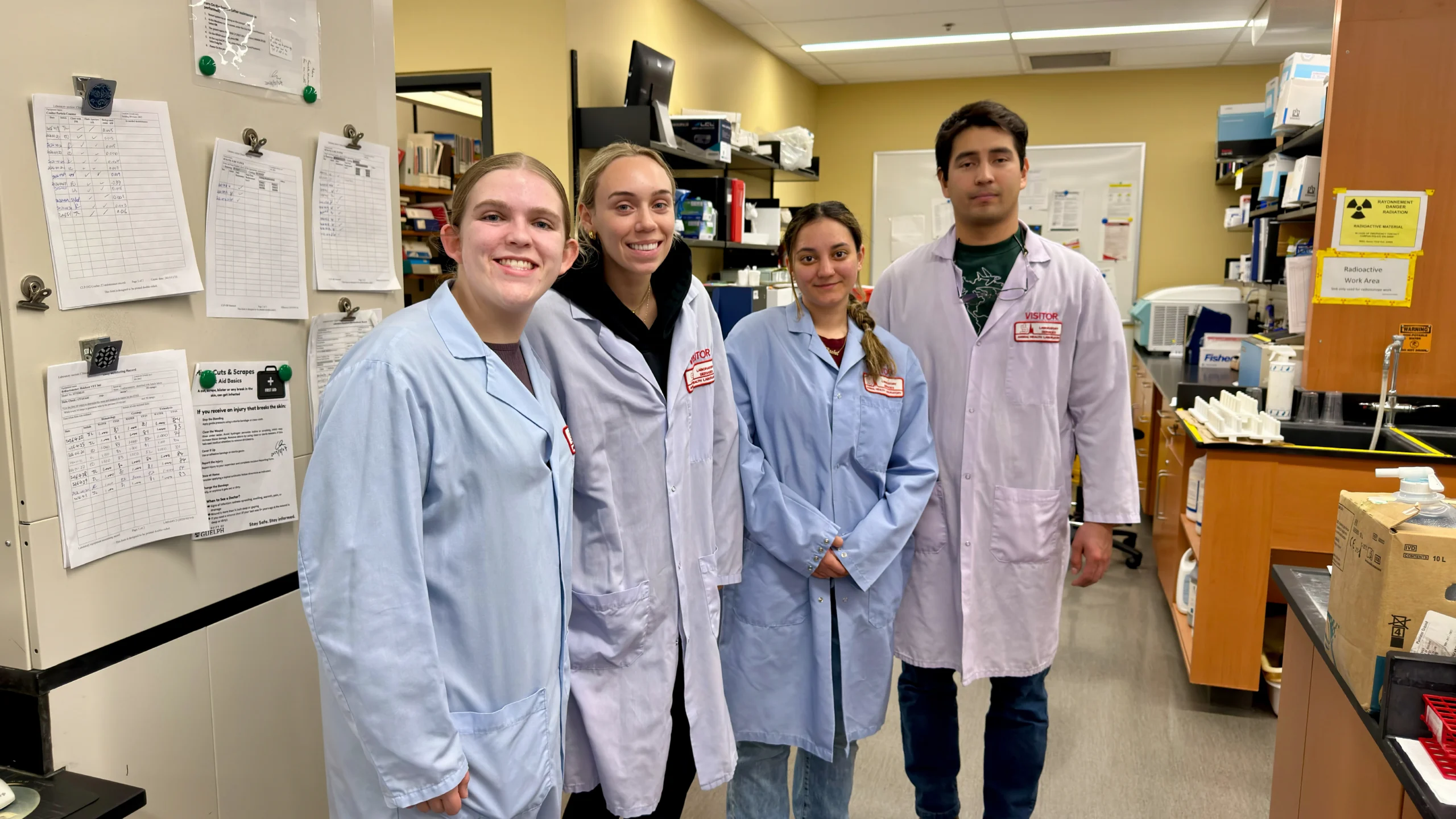 Four university researchers wearing light blue laboratory coats and visitor badges pose for a photo inside the Animal Health Lab, with research equipment and a radioactive work area visible in the background.