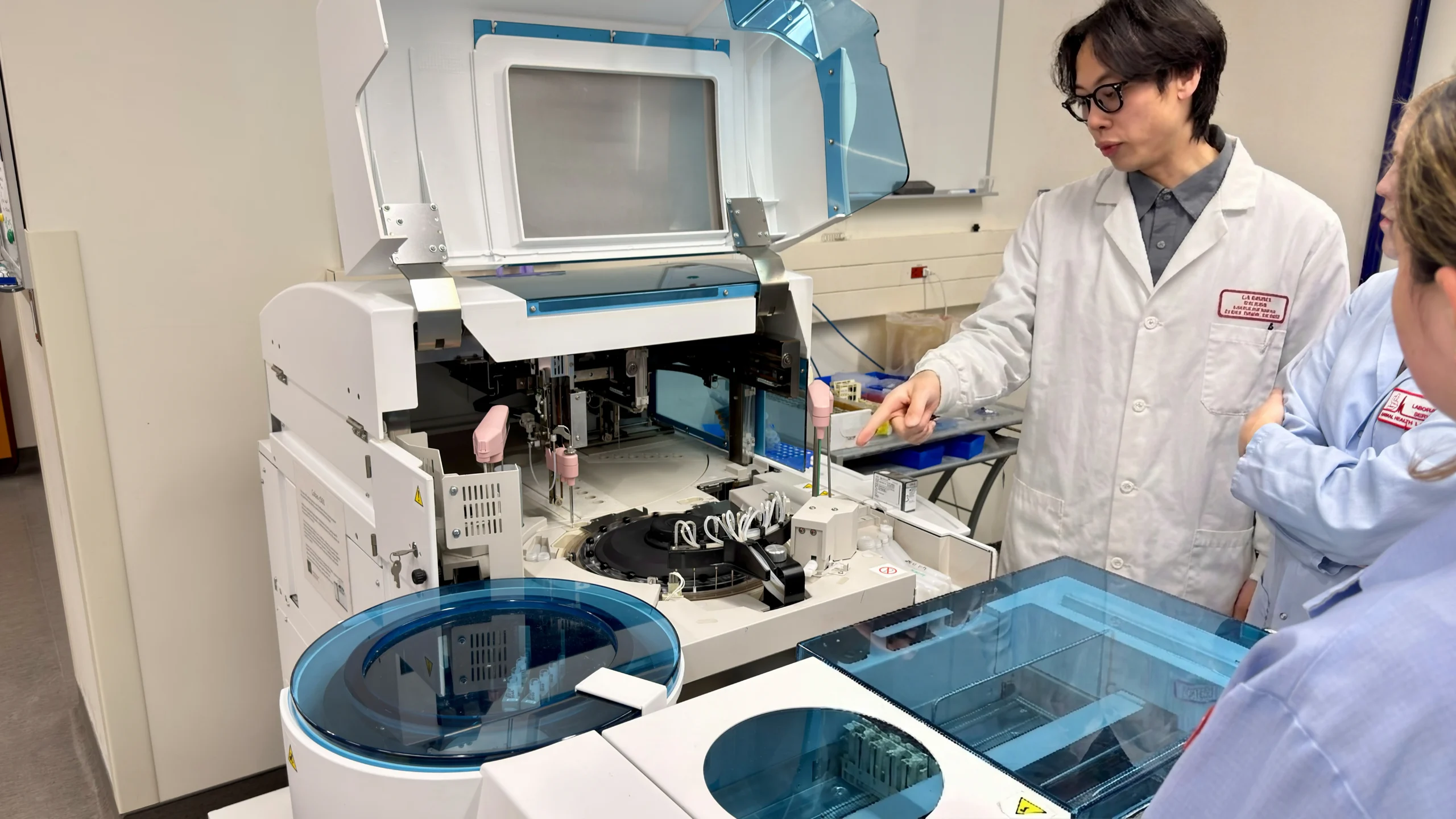 A researcher in a white lab coat points to the internal components of an open clinical chemistry analyzer while explaining its operation to colleagues during a laboratory technical visit.
