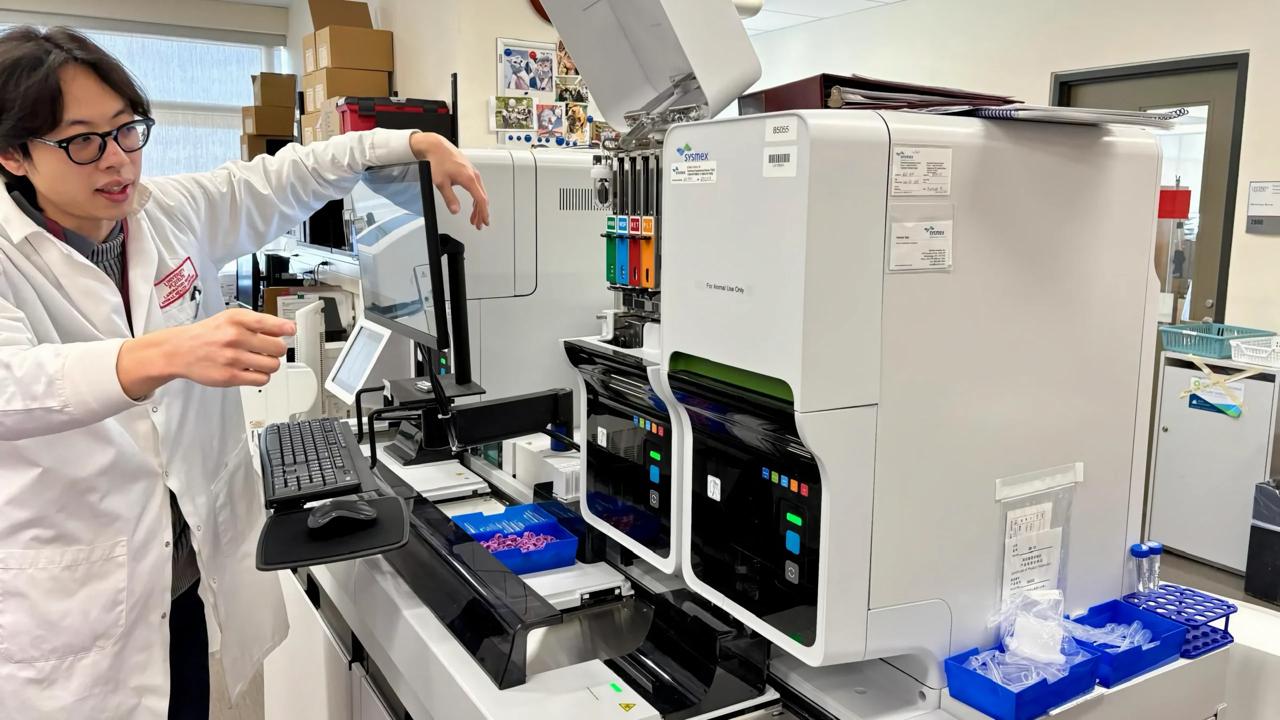 A researcher in a white lab coat demonstrates the use of a Sysmex automated hematology analyzer and its interface during a technical presentation in a diagnostic laboratory.