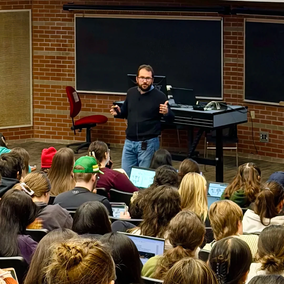 Home Dr. Ribeiro gestures while lecturing at the front of a university classroom filled with students sitting at desks with laptops.