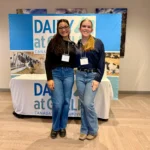 Outreach Amanda and Alicia smiling in front of a "Dairy at Guelph" display and information table during the Dairy at Guelph Research Showcase 2025.