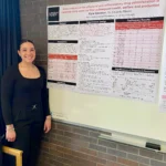 Outreach A researcher stands smiling next to a scientific poster titled "Meta-analysis of the effects of anti-inflammatory drug administration in transition dairy cows on their subsequent health, welfare and production" displayed on a whiteboard against a dark brick wall.