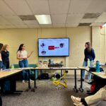 Outreach Three female researchers present a slide titled "Opportunities to Improve Streamlined Assessments" on a digital screen during a laboratory meeting with Dr. Stefan Borchardt and colleagues.