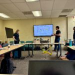 Outreach A researcher presents a slide titled "Treatment vs Prevention" on a digital screen to Dr. Stefan Borchardt and a group of students during a university laboratory meeting.