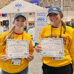 Outreach Alicia and Kylie in yellow University of Guelph sweaters and "I Care for Cattle" caps smile while holding "Dairy Diploma" certificates at the Royal Winter Fair 2025.