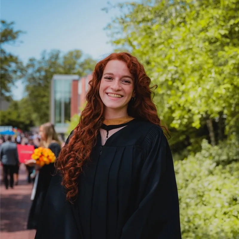 Members A smiling woman with long, curly red hair wearing a black academic graduation gown, posing outdoors on a university campus with green trees and buildings in the background.