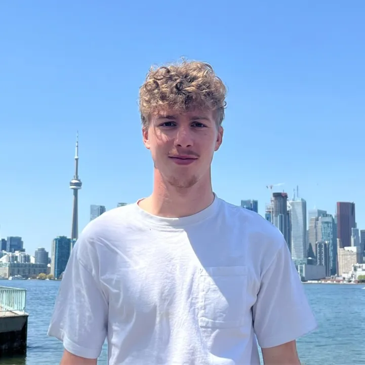 Members A portrait of a man with curly blonde hair wearing a white t-shirt, posing in front of the Toronto skyline and waterfront under a clear blue sky.