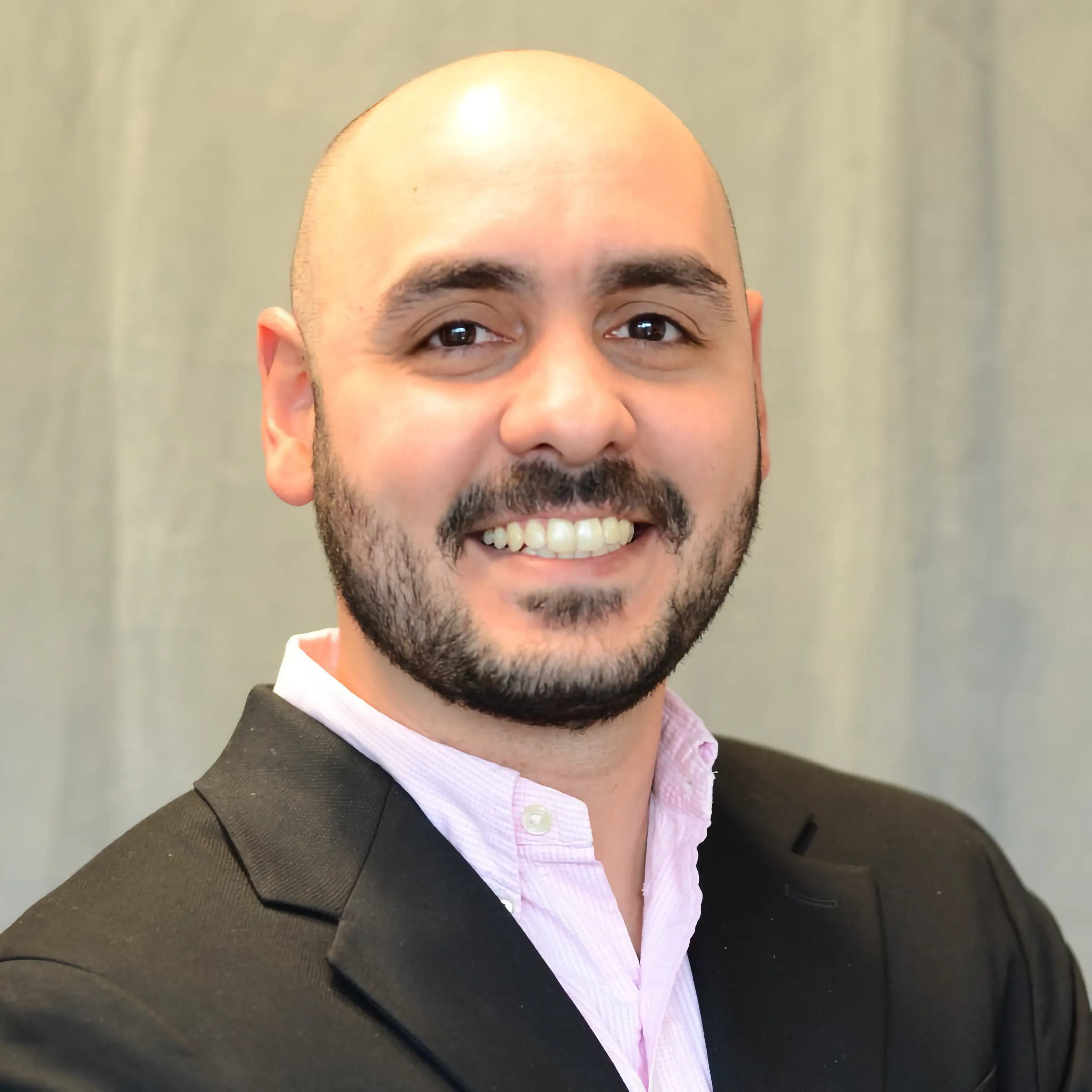 Members A portrait of a smiling bald man with a dark beard and mustache, wearing a light pink button-down shirt under a black blazer against a neutral grey background.