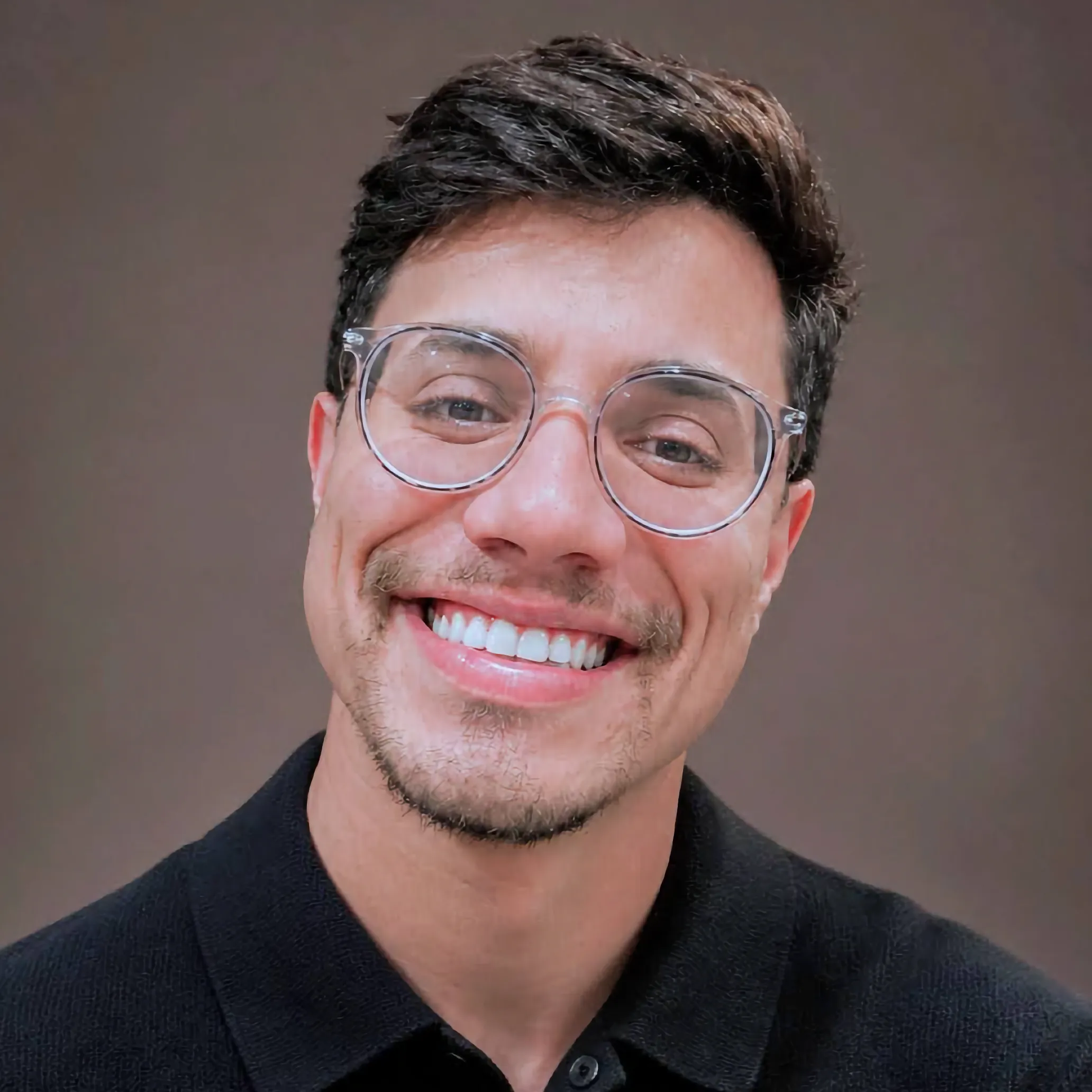Members A portrait of a smiling man with short dark hair and a light beard, wearing clear-framed round glasses and a black polo shirt against a neutral brown background.
