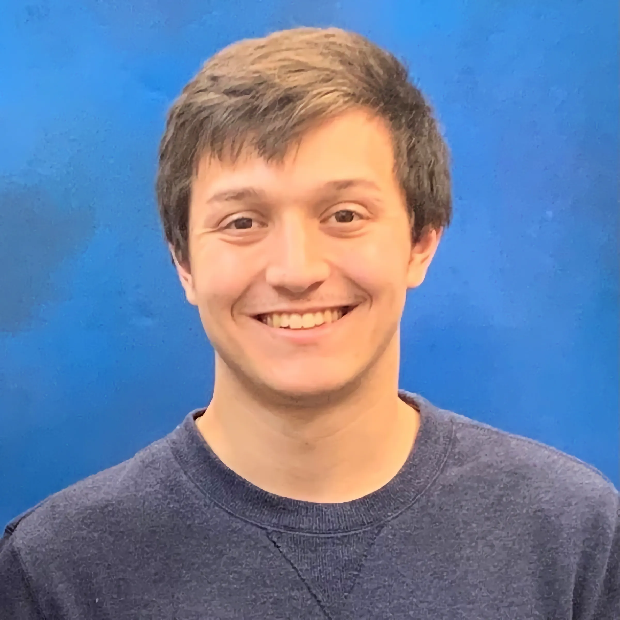 Members A portrait of a smiling man with short brown hair, wearing a navy blue crew neck sweater against a solid blue background.