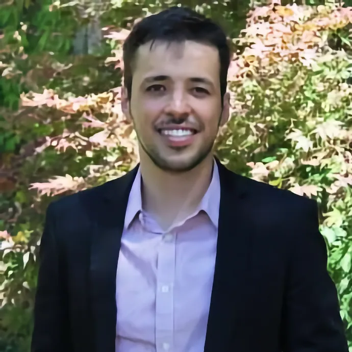 Members A portrait of a smiling man with short dark hair and a light beard, wearing a light pink button-down shirt and a black blazer, with a background of green and reddish foliage.