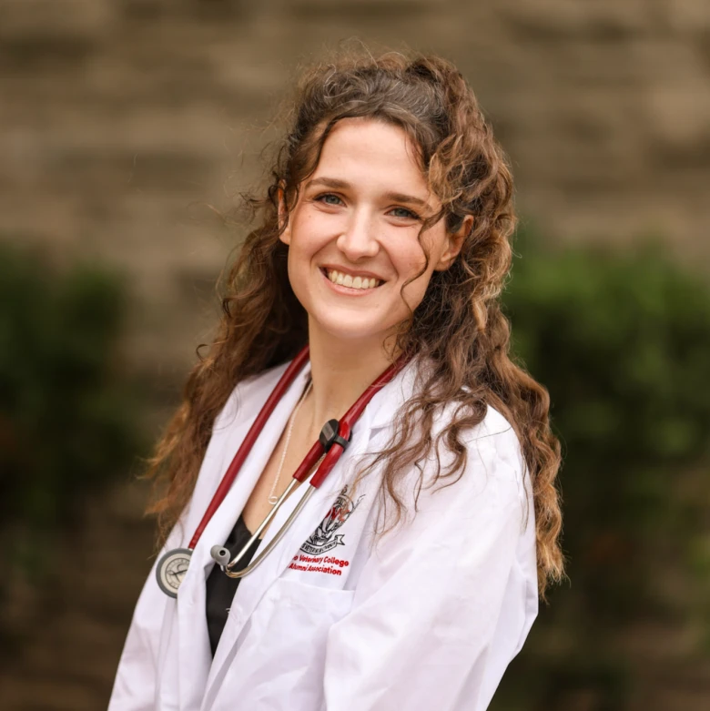 Members Professional portrait of a smiling woman with long wavy brown hair wearing a white lab coat with an institutional crest and a red stethoscope around her neck, outdoors.