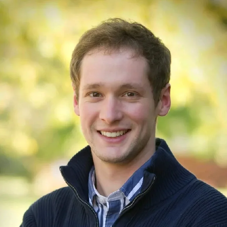 Members A professional headshot of a smiling male researcher wearing a dark zip-up sweater and a collared shirt, with a blurred natural green background.