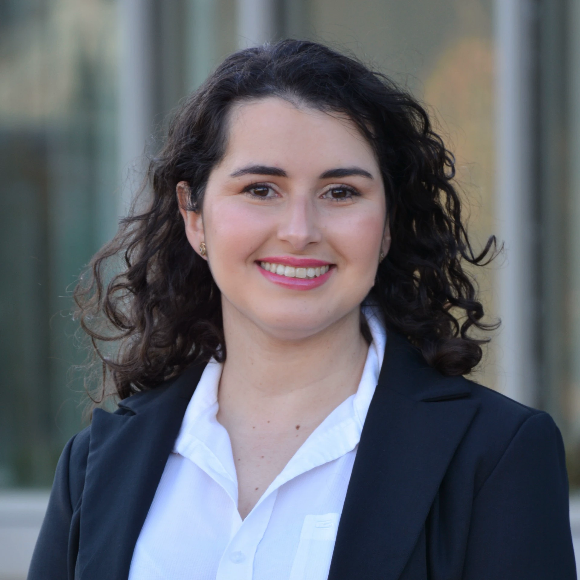 Members Professional portrait of a smiling woman with long curly dark hair wearing a white collared shirt and a black blazer, standing outdoors with a blurred building facade in the background.