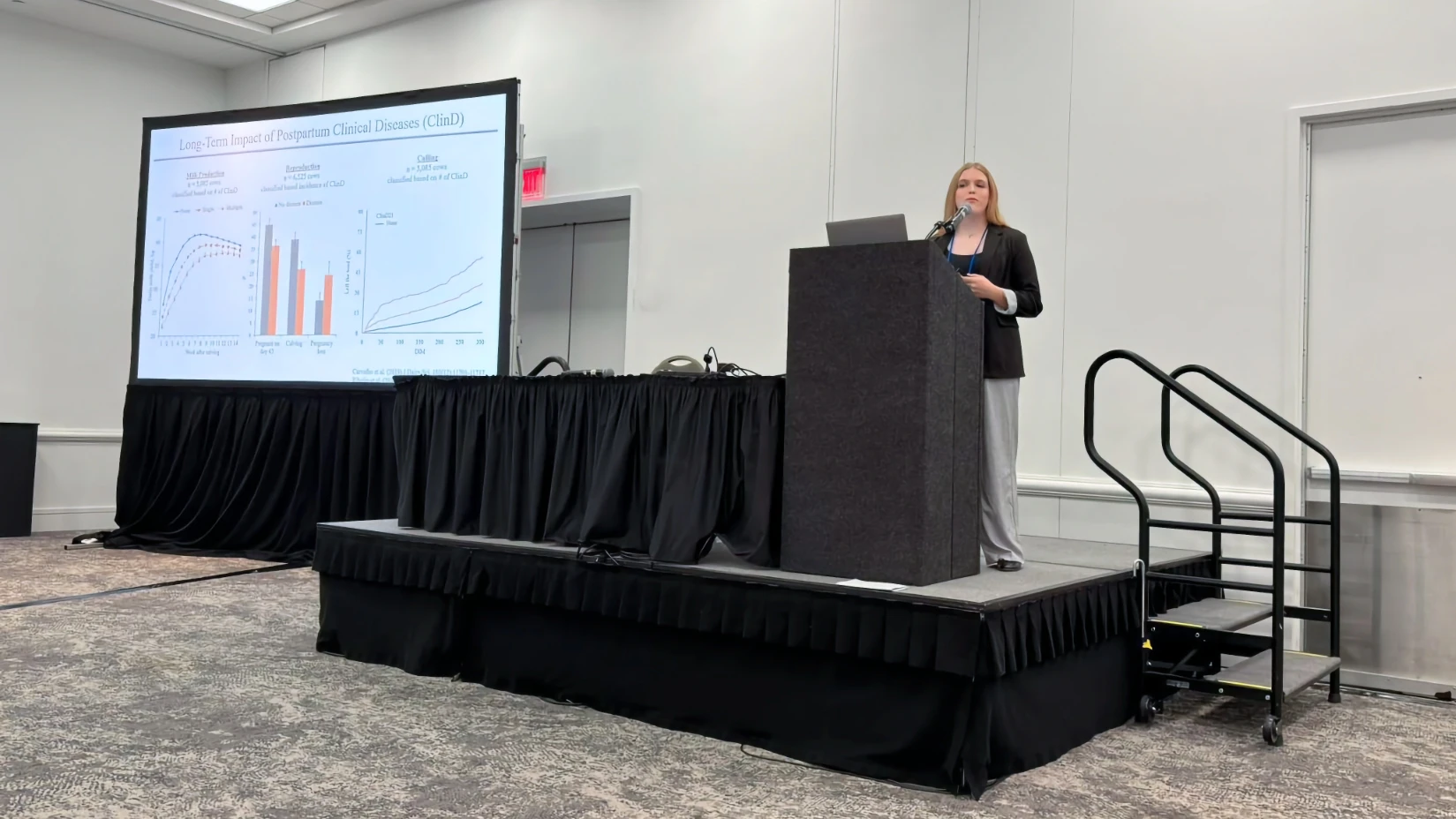 Ribeiro Lab at ADSA 2026 Wrap-Up A female researcher giving an oral presentation at a conference podium. The large screen behind her displays a slide titled 'Long-Term Impact of Postpartum Clinical Diseases (ClinD)' featuring multiple statistical line graphs and bar charts plotting milk production, reproduction, and culling data.
