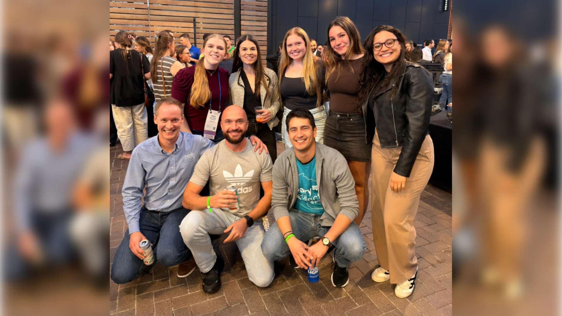Ribeiro Lab at ADSA 2026 Wrap-Up A group of eight researchers smiling and posing together at an evening social event during a conference. Three individuals are kneeling in the front row, and five are standing behind them against a modern wood-paneled and dark metal architectural wall decorated with string lights.
