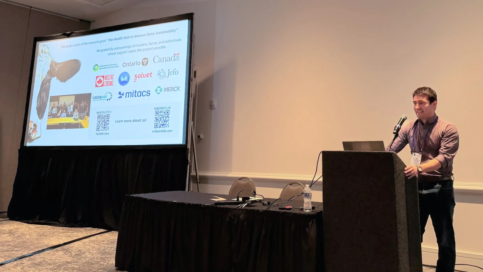 Ribeiro Lab at ADSA 2026 Wrap-Up A male researcher presenting at a podium microphone during a scientific conference. The slide on the large projection screen behind him acknowledges research grants and partners, displaying institutional logos including the University of Guelph, Ontario, Canada, NSERC, Mitacs, Merck, and the Ribeiro Lab.