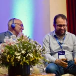 Outreach Two male speakers with microphones sitting on a couch at a conference stage, next to a flower arrangement. The person on the right is looking down and adjusting their equipment, and the one on the left is wearing a translation headset.