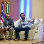 Outreach Two speakers with microphones presenting from a couch at an event. The person on the right is speaking and gesturing, and the one on the left is listening while wearing a translation headset. National flags are visible in the background.