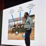 Outreach A speaker on a stage pointing with their right hand to specific points on a large presentation screen. The screen shows names and corresponding geographic locations in Argentina (Corrientes, Buenos Aires, Cordoba) connected by arrows to photos. To the left is an event banner with the CONIL logo and a cow illustration.
