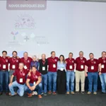 Outreach A group of about twenty people in matching burgundy polo shirts and jeans posing for a photo on a stage. Most are standing, with two kneeling in the front. They are in front of a screen displaying the text 'NOVOS ENFOQUES 2026' and the logos of 'CONAPEC Jr.' and 'Texas A&M AGRILIFE RESEARCH'.