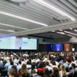 Outreach A view from the back of a large auditorium filled with people attending a conference. A speaker is visible on the distant stage presenting in front of two large screens. The screens show text with graphs and tables under the heading 'NOVOS ENFOQUES 2026' and the title "Timing of Embryo Transfer or Removal Affect Luteal Lifespan in Sheep".