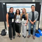 Outreach Four researchers posing together in front of a scientific poster presentation at an academic conference hall. The research poster displays University of Guelph branding and is titled 'Gene expression signatures in prepartum peripheral blood leukocytes associated with postpartum health status in dairy cows'.