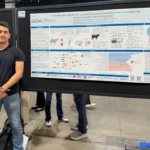 Outreach A male researcher standing with crossed hands beside a large research poster display at a scientific event. The poster outlines a study on gene expression signatures in prepartum peripheral blood leukocytes and dairy cow health, featuring a prominent schematic diagram and data charts.