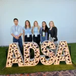 Outreach Five laboratory members standing together behind large, lighted marquee letters spelling 'ADSA' with a cow-print pattern pattern, set on a section of artificial grass during a conference.