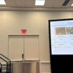 Outreach A female researcher speaking from a podium during an oral presentation at a conference. Beside her, a large projection screen displays a slide titled 'Expanding the Data Set,' containing a data table and photos labeled from different dairy farms.